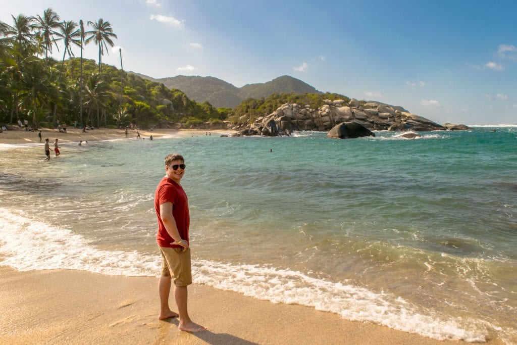 身穿红色衬衫的杰里米·斯托姆站在塔罗纳国家公园卡波圣胡安海滩的水边。我们绝对推荐您在哥伦比亚14天的行程中参观这个海滩!