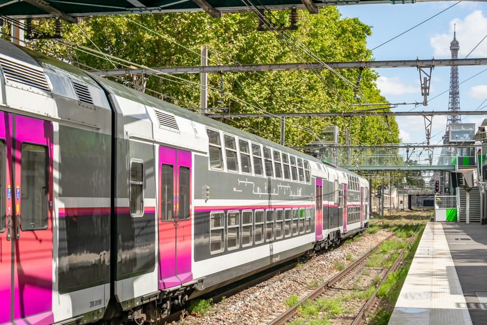 Photo of a pink and white train in a station in Paris. You can see the Eiffel Tower in the top right of the photo. If you follow this 3 day Paris itinerary, you might take this train to Versailles.