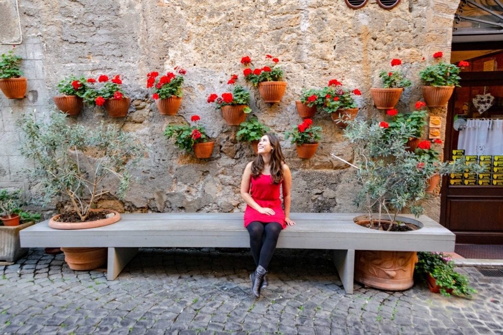 凯特坐在意大利奥维多的长凳上。她穿着一件红裙子，黑色紧身衣和黑色靴子。她身后墙上的花盆里有红花。
