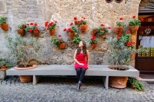 凯特·斯道姆坐在意大利奥维托的长凳上。她穿着一件红裙子，黑色紧身衣和黑色靴子。她身后墙上的花盆里有红花。