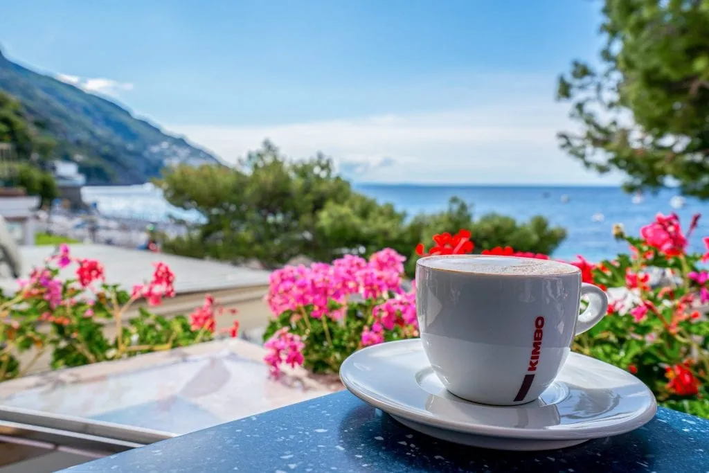 粉红色和红色的花朵与波西塔诺海滩的背景和一杯咖啡的白色碟子在前景。