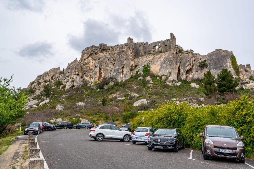 Parking lot outside of Les Baux-de-Provence. Several cars are parked to the right and the city is visible in the background.