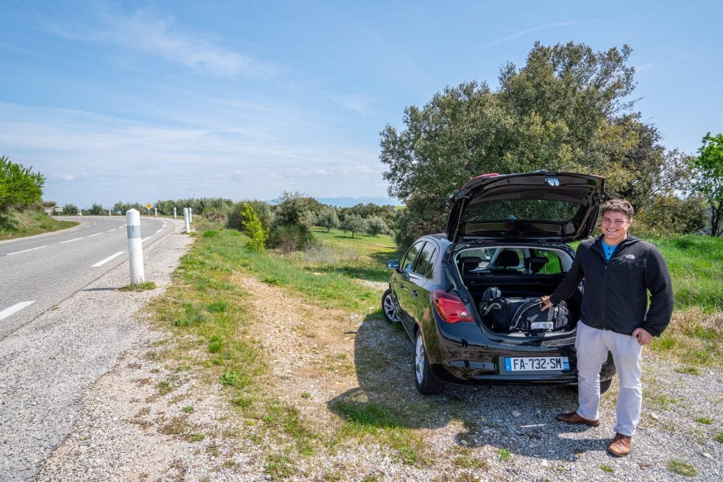 在我们的法国自驾游中，杰里米站在乡村公路的右边。他站在一辆黑色租来的车前，后面的舱门是开着的，他穿着一件黑色夹克。