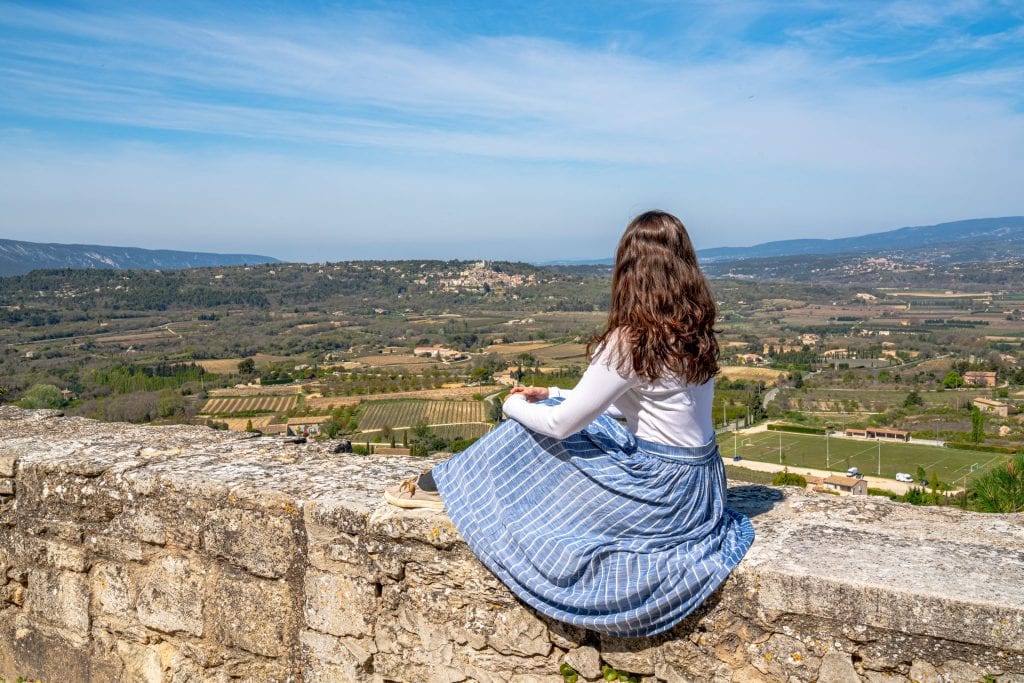 凯特穿着蓝色裙子坐在石墙上俯瞰吕贝隆山谷的乡村。摄于法国博尼埃。