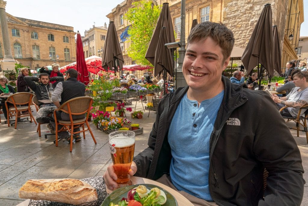 Jeremy in a blue shirt and black jacket, holding a beer while eating lunch in a square of Aix-en-Provence