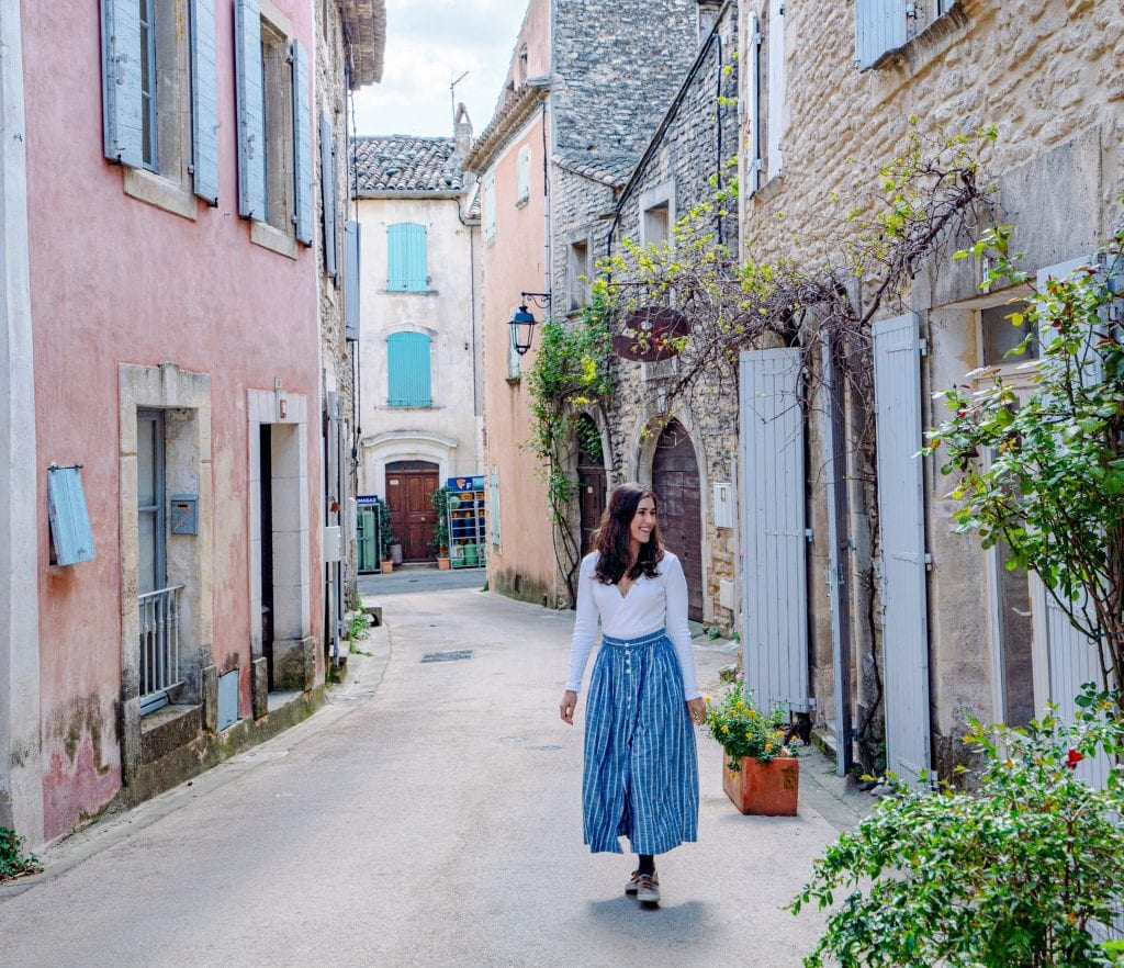 凯特在我们的法国自驾游中走在古尔特一条狭窄的街道上。在她的左边有一座粉红色的建筑，她穿着一条蓝色的长裙。