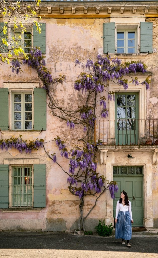 Kate in a long blue skirt standing in front of a building in Goult with green shutters. Wisteria is blooming on the building. Don't miss visiting Goult during your south of France itinerary!