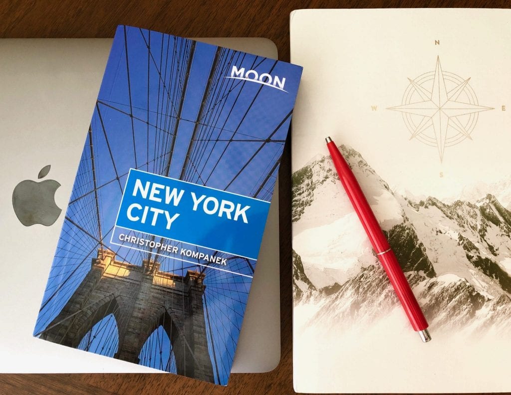 Macbook Pro的照片，一个封面上有山的笔记本和一支红色的钢笔。一本《月亮纽约》放在它们上面——用它来找到一些在纽约中城最好的事情!