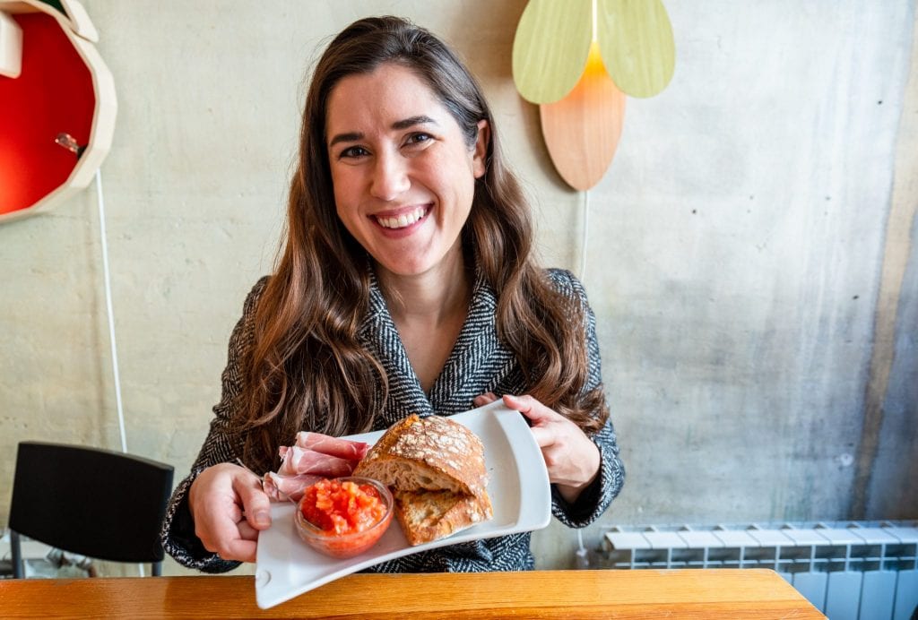 身着灰色外套的Kate Storm端着一盘面包、西红柿和火腿——el b_us是你在马德里3天行程中吃早餐的好去处!