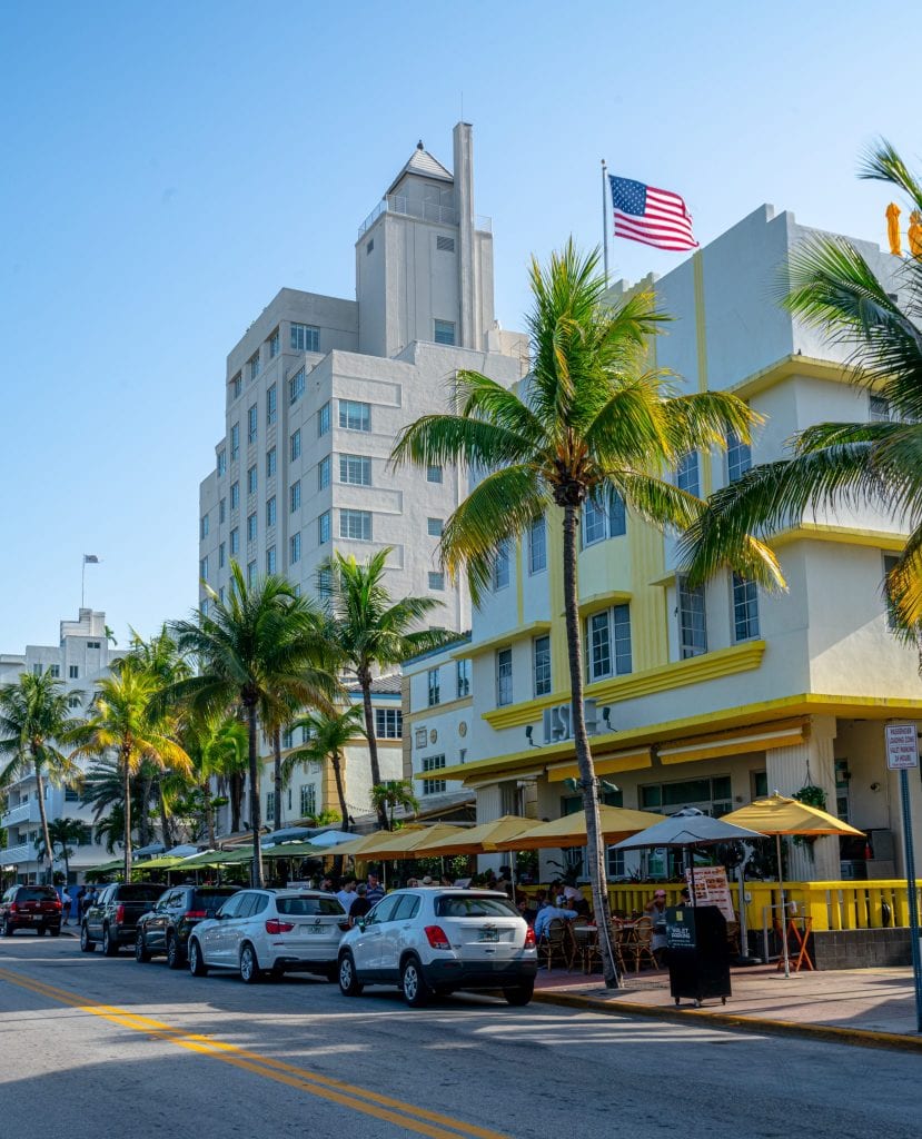 迈阿密海滩的海洋大道，迈阿密之旅的必看景点!街道两旁种着棕榈树，前景中有一座黄白相间的建筑。照片上方飘扬着一面美国国旗。