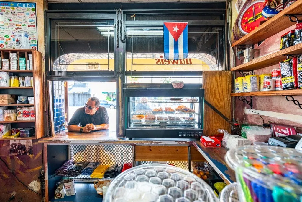 站在小哈瓦那El Pub窗口等咖啡的男子。右边是一盒肉馅卷饼，照片的右上角附近悬挂着一面古巴国旗。