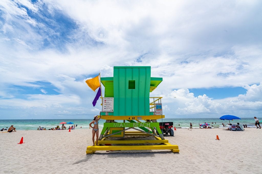 凯特站在南海滩的绿色装饰艺术救生员站上——这是迈阿密3天行程中必不可少的地方!救生员站写着“迈阿密海滩”，上面有两面旗帜。