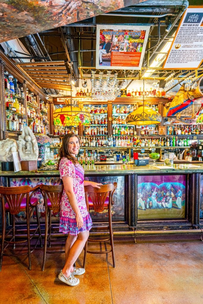 凯特身穿粉色连衣裙站在Cubaocho，站在一个历史悠久的木制酒吧前，酒吧里有许多玻璃瓶和吊灯。你在迈阿密的长周末可别错过这个!