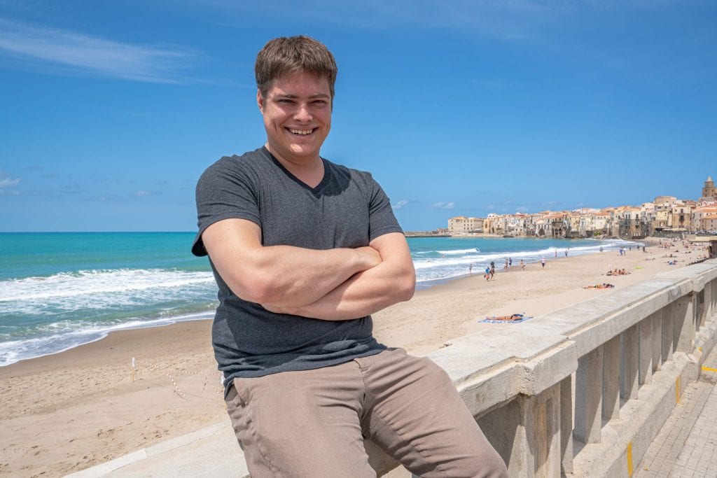 杰里米·斯托姆坐在俯瞰塞法卢海滩的墙上，这是西西里岛公路旅行行程中的一个神话般的站点