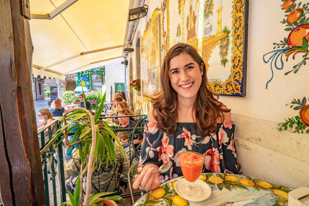 凯特·斯道姆坐在陶尔米纳BamBar餐厅的桌子上。她面前有一个橙色的格兰尼塔，她身后有黄色的瓷砖。参观BamBar绝对是西西里岛陶尔米纳最好的事情之一!