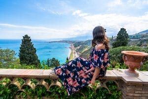 在西西里岛陶尔米纳，凯特·斯道姆身穿碎花连衣裙坐在花园墙上俯瞰大海