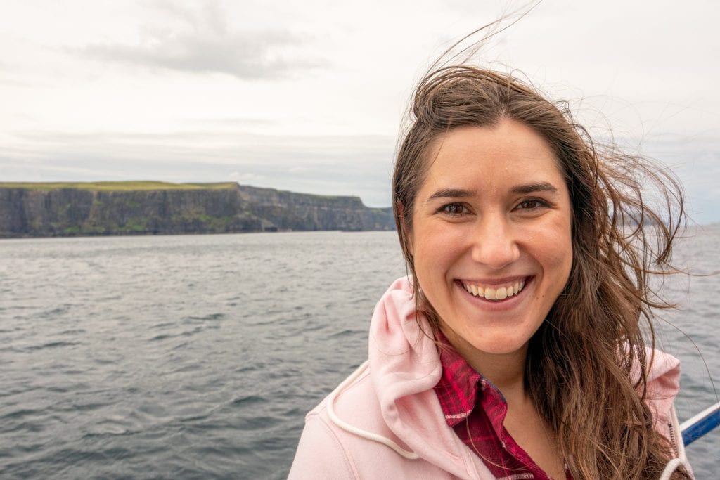 凯特·斯道姆身穿粉色夹克，在莫赫悬崖巡航时对着镜头微笑——悬崖在背景中可见。