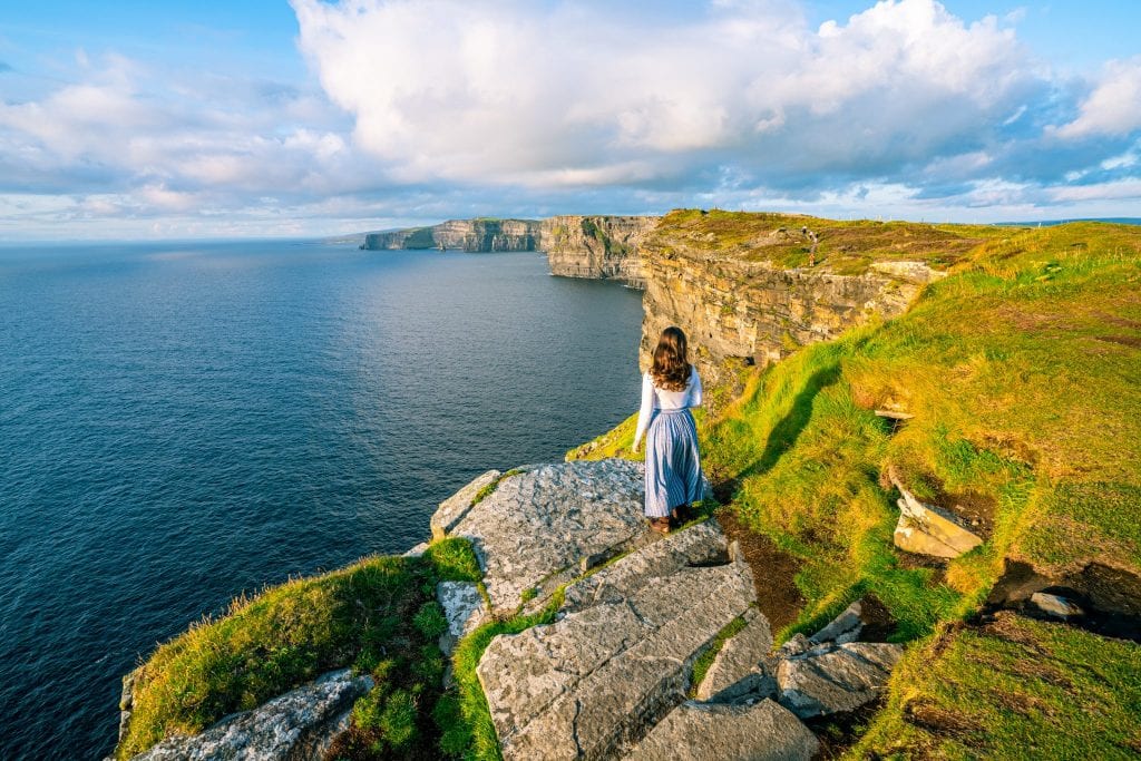 凯特·斯道姆穿着蓝色裙子站在爱尔兰莫赫悬崖前。她把目光从镜头上移开了。