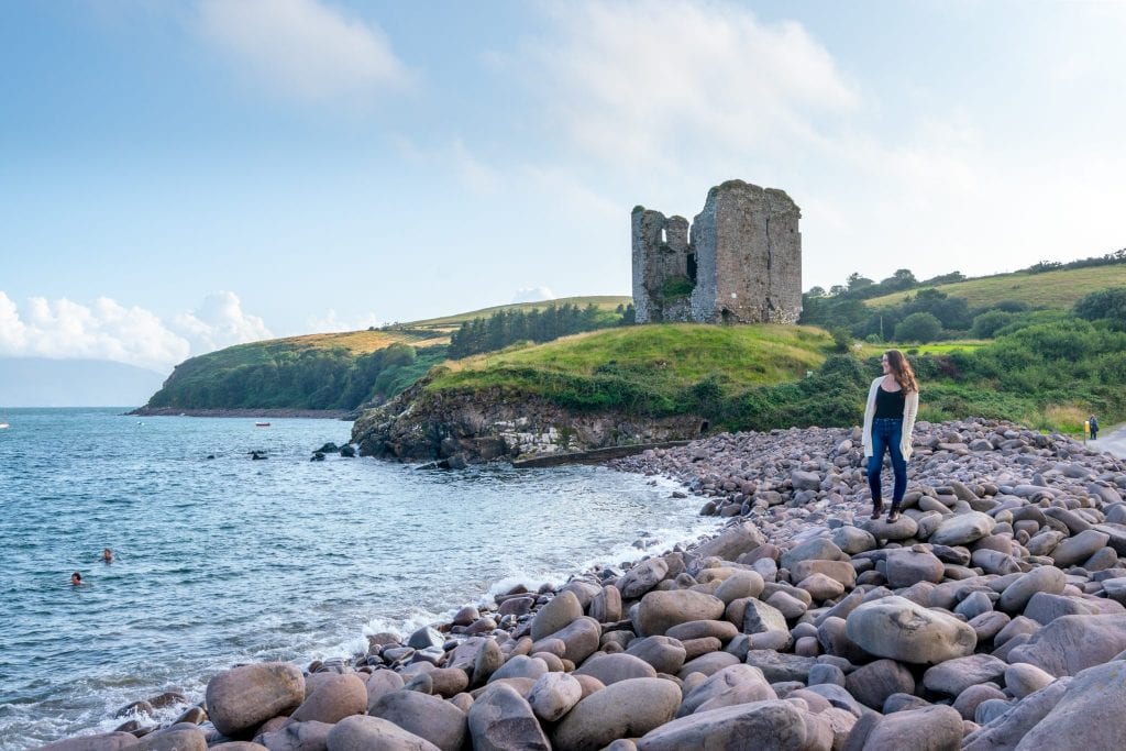 凯特·斯道姆站在爱尔兰丁格尔半岛的卵石海滩上。密纳德城堡在她身后。这是爱尔兰着装的一个很好的例子!
