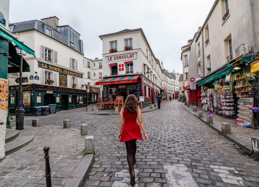 凯特·斯道姆身着红色连衣裙站在蒙马特区的La Consulat咖啡馆前，这是巴黎最上镜的地方之一