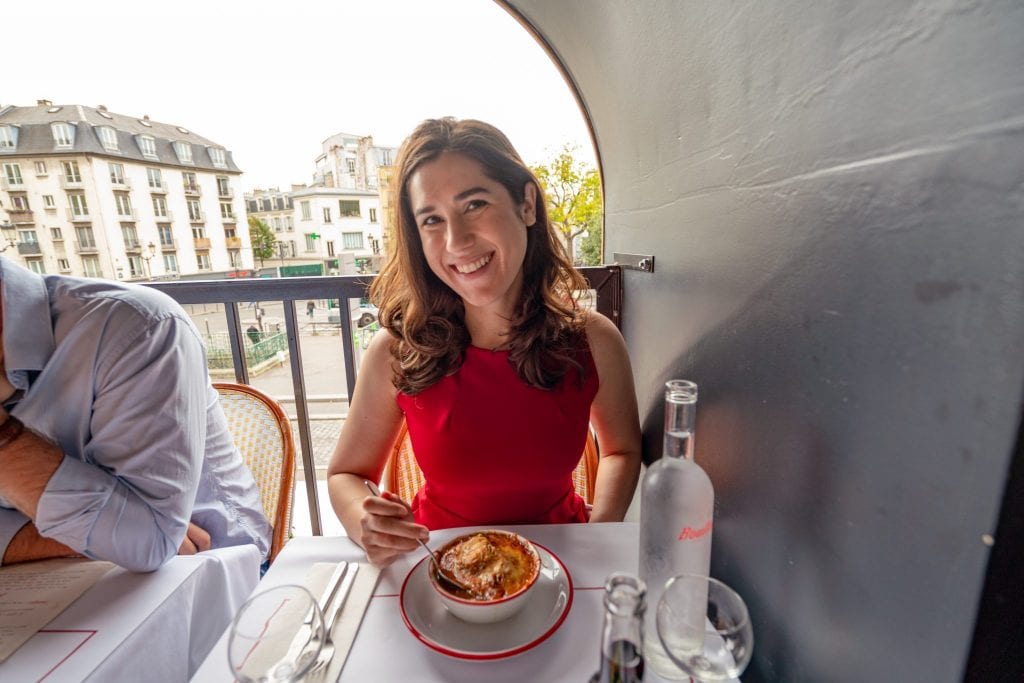 凯特·斯道姆身穿红色连衣裙，面前的桌子上放着一碗法式洋葱汤——猪肉汤是你周末巴黎之旅的午餐好选择!