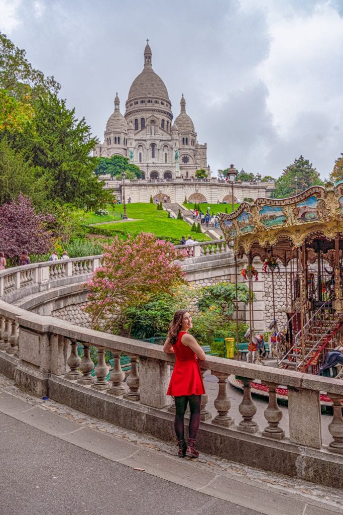 Kate Storm in a red dress standing with a caroseul and Sacre Coeur in the background--this is one of the most instagrammable places in Paris!