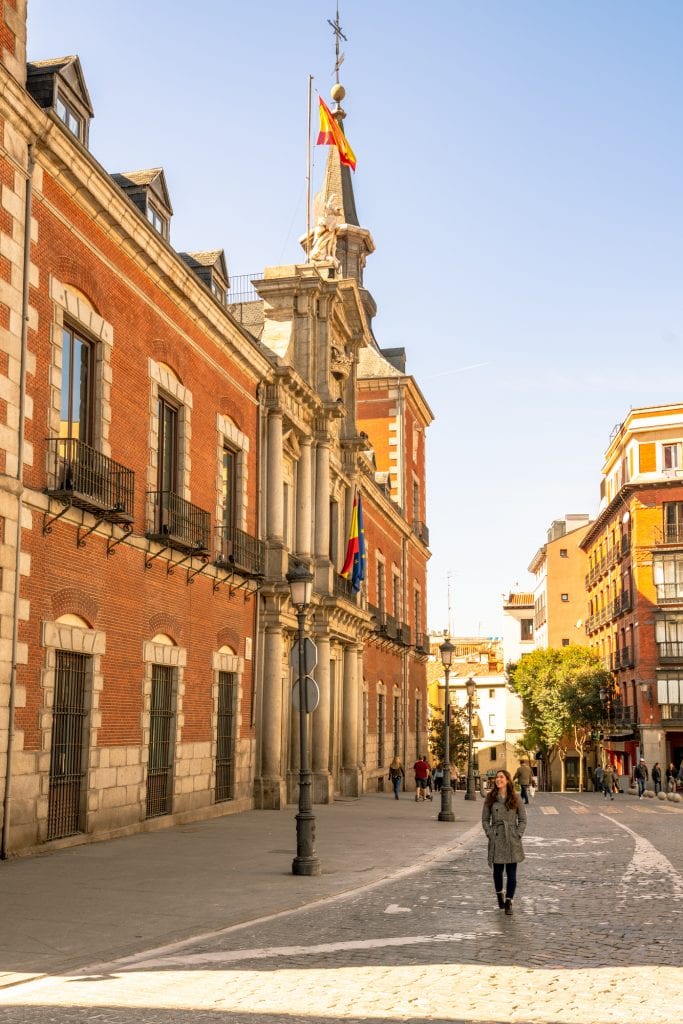 身着灰色外套的凯特·斯道姆走在马德里的街道上，左边是一座红砖建筑