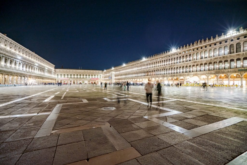 威尼斯圣马可广场夜晚的照片，几个人在广场上走来走去，建筑物上的灯光照亮了广场