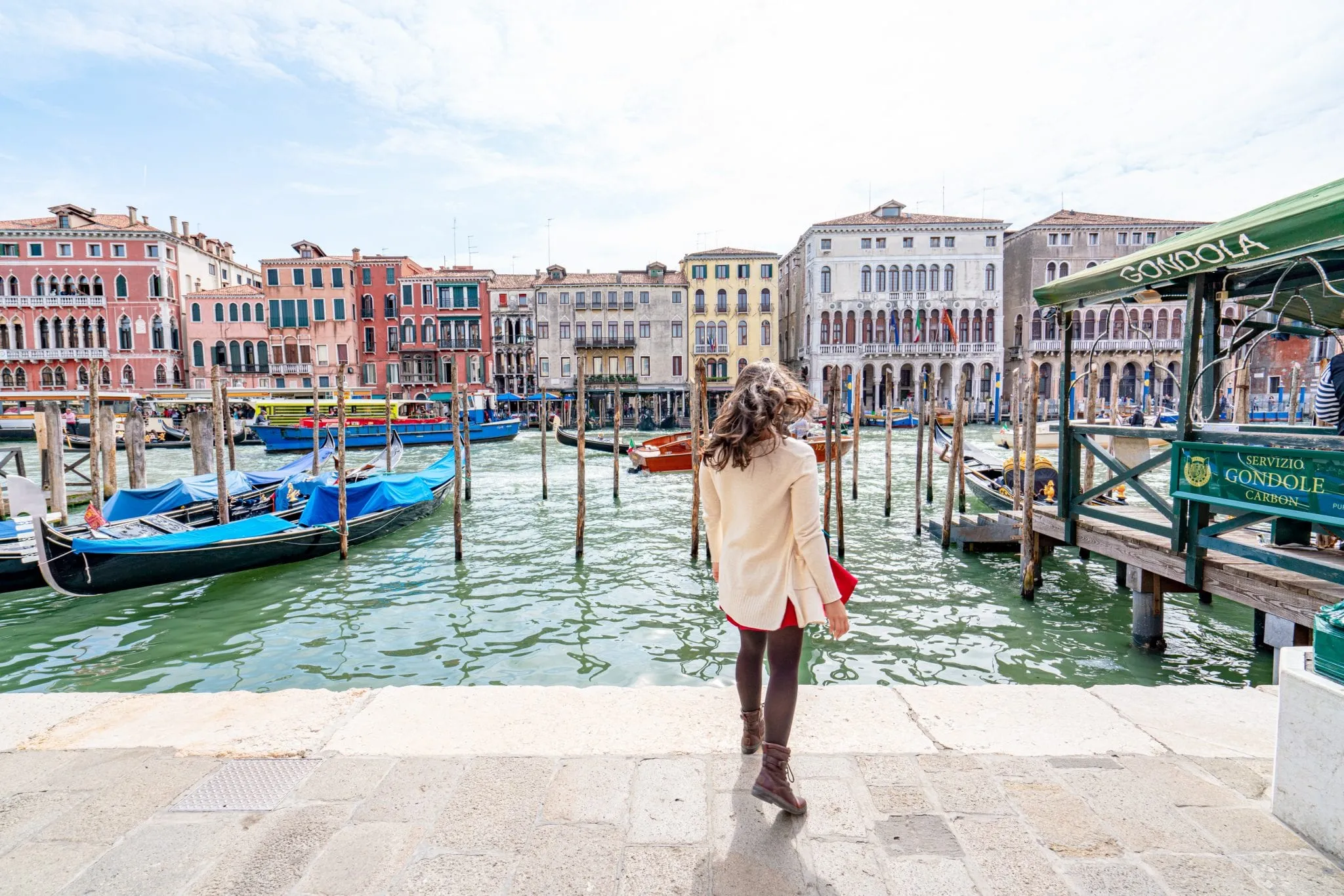 凯特·斯道姆背对着镜头站在大运河上，这是佛罗伦萨到威尼斯一日游的必看景点!贡多拉停在运河边，凯特穿着一件奶油色的毛衣。