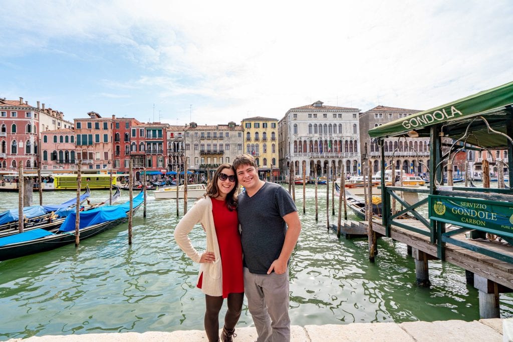 凯特·斯道姆和杰里米·斯道姆站在威尼斯大运河边。凯特穿着一件红裙子，身后是贡多拉。