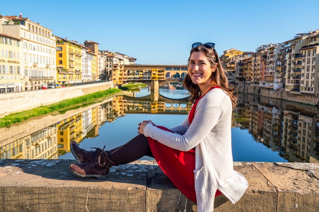 身着红裙的凯特·斯道姆坐在阿尔诺河前的一座桥上，背景中可见维奇奥桥——在意大利佛罗伦萨的2天里，一定要看看这幅风景