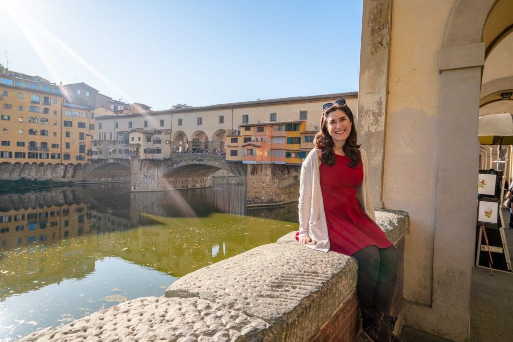 凯特·斯道姆穿着红裙子坐在墙边，背景是维奇奥桥——一旦你从罗马开车到佛罗伦萨，这些风景都在等着你