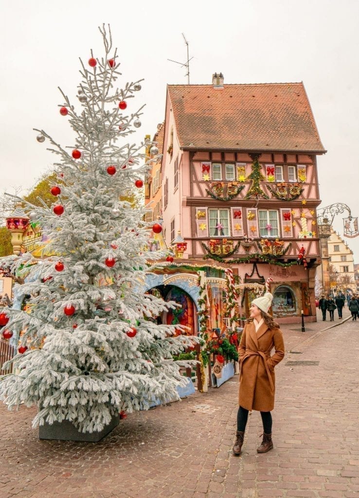 身着棕色外套的凯特·斯道姆站在科尔马的一棵白色圣诞树旁。背景是一个装饰过的粉色房子——这是科尔马的一个经典的冬季拍照地点!