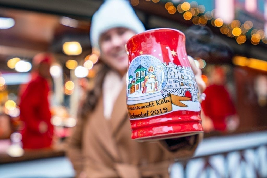 凯特·斯道姆(Kate Storm)在科隆圣诞市场拿着一个红色马克杯对着镜头，这里有欧洲最好的圣诞市场