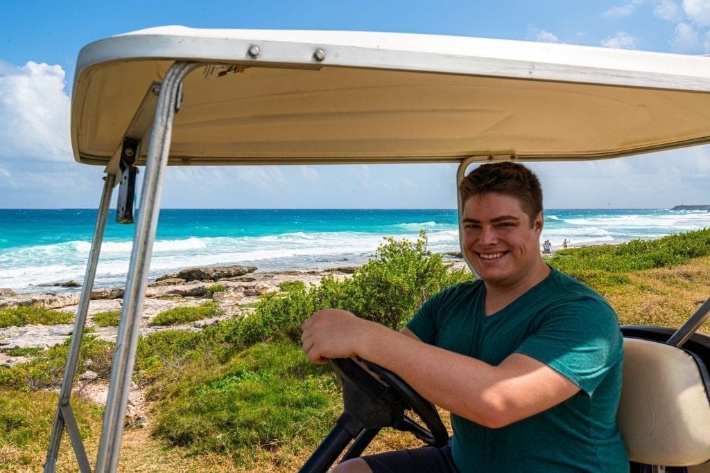 身穿绿色衬衫的杰里米·斯托姆坐在高尔夫球车的方向盘后面，背景是Isla Mujeres的东海岸。租一辆高尔夫球车是在Isla Mujeres最好的事情之一