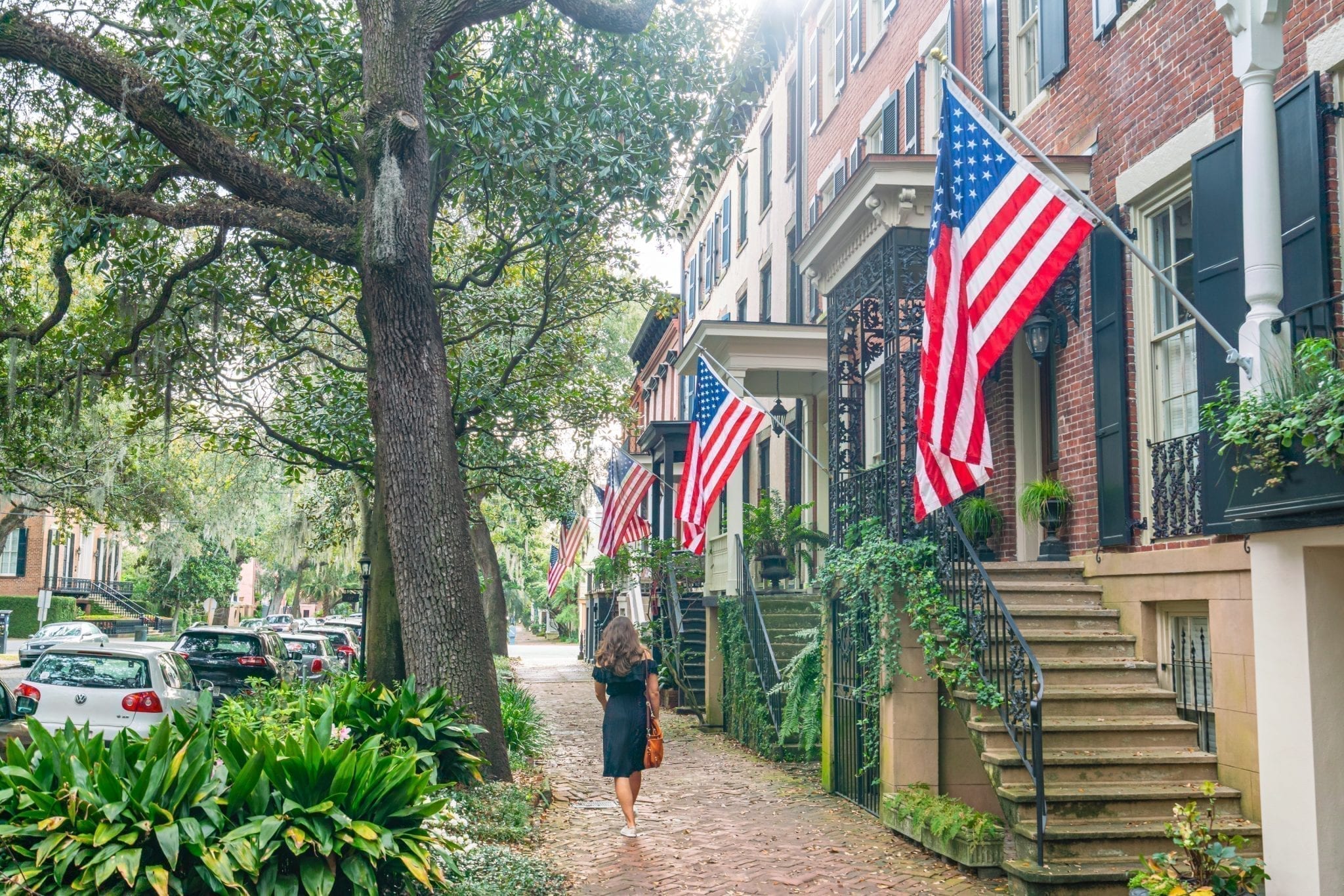 凯特·斯道姆身穿黑色长裙，沿着佐治亚州萨凡纳的琼斯街从镜头前走开，照片的右侧是几面悬挂在房屋上的美国国旗