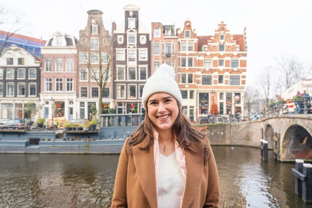 在一个多云的日子里，身穿棕色外套、头戴白色帽子的凯特·斯道姆站在阿姆斯特丹的一条运河前