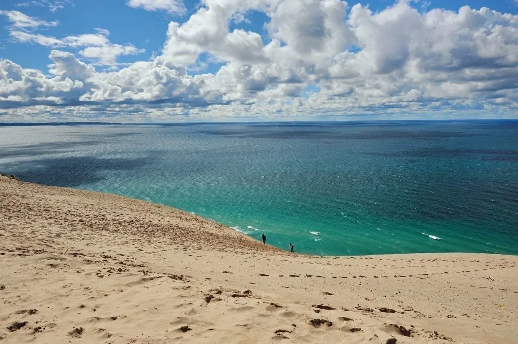 密歇根湖的沙丘，背景是蓝绿色的湖，远处有两个小人。密歇根湖是美国一些最好的海滩的所在地