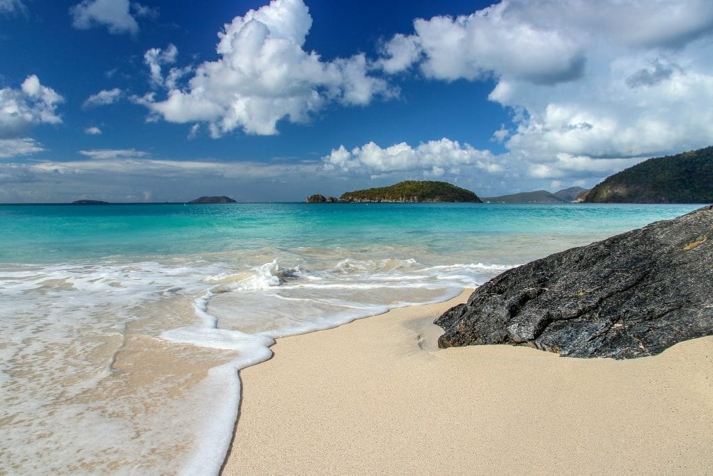 阳光明媚的日子里，圣约翰肉桂湾亮蓝色的海水，前景是金色的沙子，照片右侧是黑色的岩石。这个加勒比海滩真美。