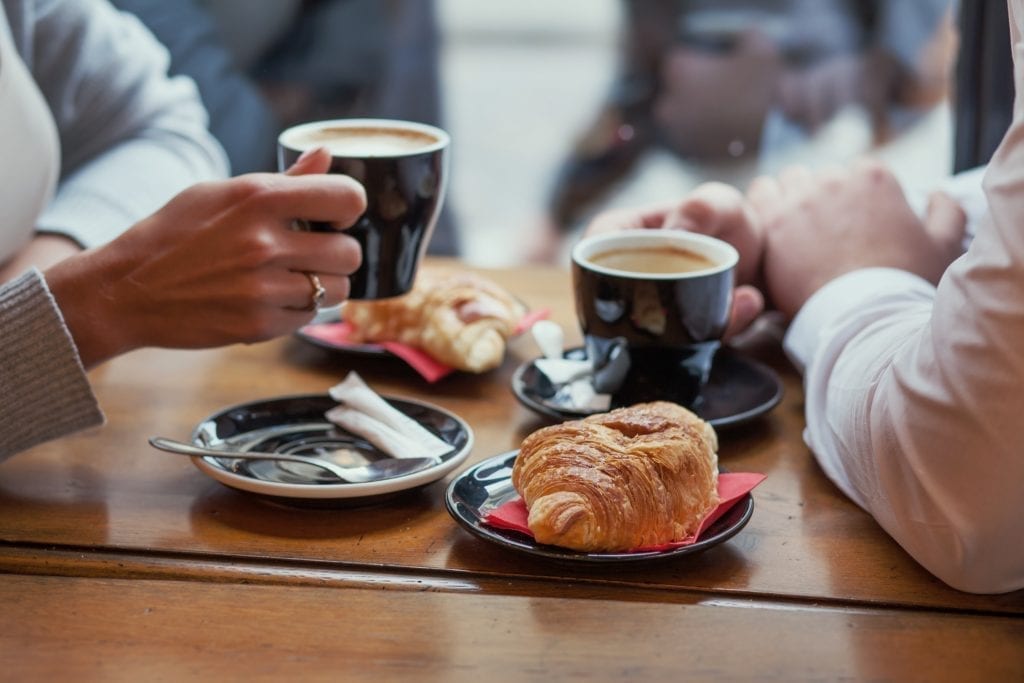 在法国，两个人拿着咖啡杯，放在一张棕色的桌子上，两边放着牛角面包。图像的焦点是食物。