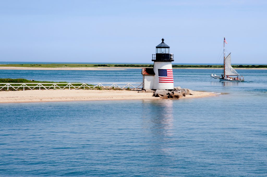 马萨诸塞州楠塔基特岛附近的白色灯塔上有一面美国国旗和一艘帆船，这是美国最好的旅游景点之一