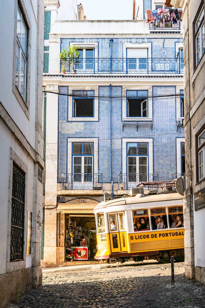 view of a yellow tram crossing in front of a tiled building from an alfama lisbon side street