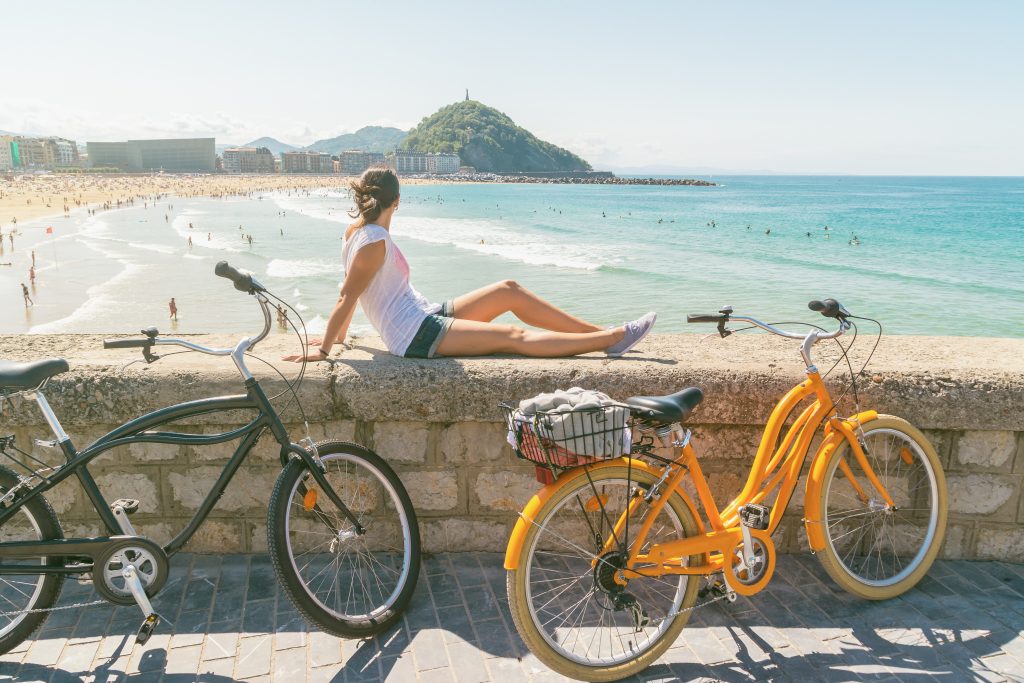 一名年轻女子坐在一堵石墙上俯瞰西班牙圣塞巴斯蒂安的海滩，前景是自行车